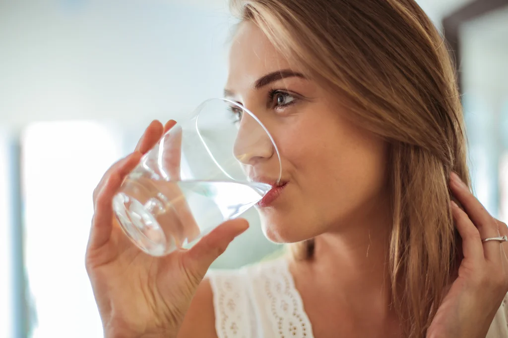 Eine blonde Frau trinkt Wasser aus einem Glas. Sie trägt ein weißes Oberteil mit Lochmuster und hat halb hochgestecktes Haar. Der Fokus liegt auf ihrem Gesicht und dem Glas. Hintergrund ist unscharf.